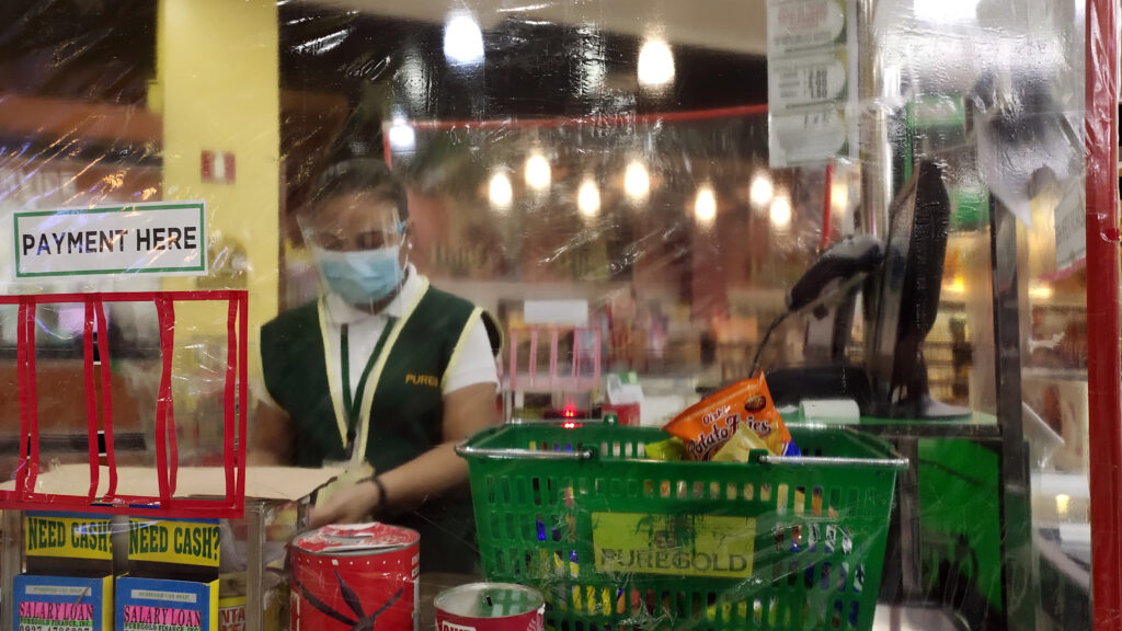 Supermarket worker is scanning groceries, they are wearing a medical mask and visor and are behind a screen. The photo was taken during the COVID-19 pandemic.