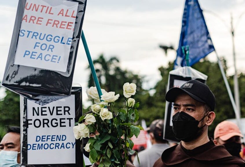 A protestor wearing a medical mask stands with two protest signs and a bunch of white roses