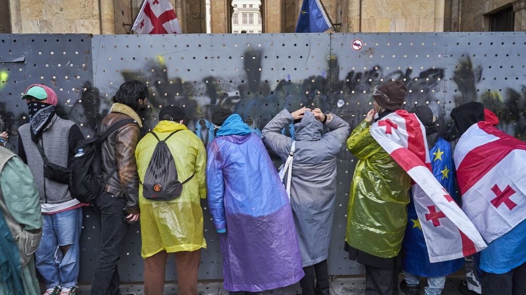 A line of protestors dressed in waterproof clothing, and some draped in Georgian and EU flags peep through an iron barrier wall.